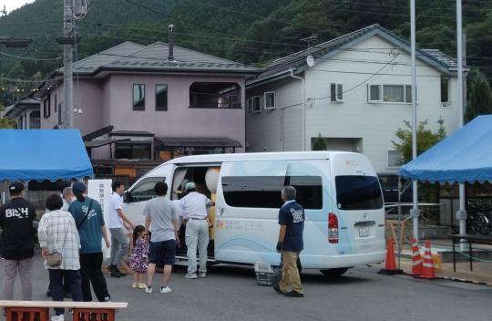 青いテントが両脇にあり、係の人が白いマルチタスク車両の中に並んでる人を誘導している写真