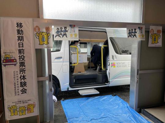 「移動期日前投票所体験会」と書かれた看板の奥に後部席のスライドドアが開いて停車しているマルチタスク車両の写真