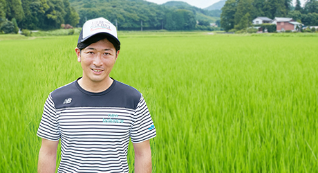 帽子を被った福田さんの後ろには鮮やかな緑の稲が植えられた田んぼが広がっている写真