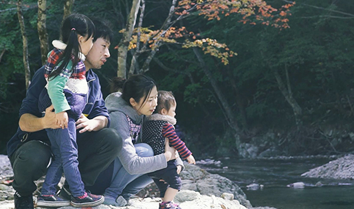川べりでお父さんとお母さんがしゃがみ込み子供2人を抱えて川の流れを眺めている写真
