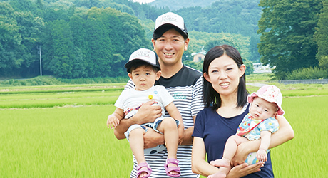 後方に広がる田んぼを背景に、夫婦2人がそれぞれ子供を抱いている福田さん家族の写真