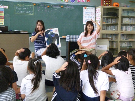女性の先生と英語指導助手が黒板の前に立ちジェスチャーをし、子ども達が左手を頭に乗せ真似をしている授業風景の写真