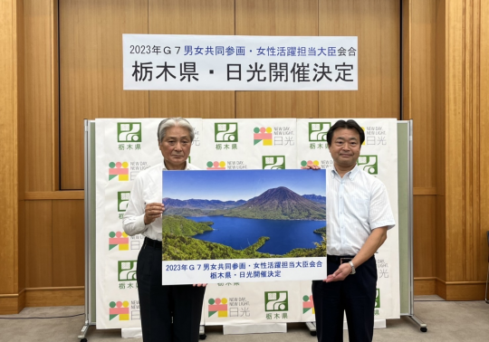 栃木県と日光市のバックボードの前で2人の男性が「2023年G7男女共同参画・女性活躍担当大臣会合 栃木県・日光市開催決定」と書かれたボードを一緒に持ち、記念撮影をしている写真