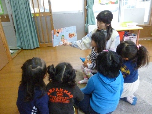 床に座り右手で絵本を持ち膝に女の子を抱きながら読み聞かせをしている先生と、床に座って聞いている子ども達の写真