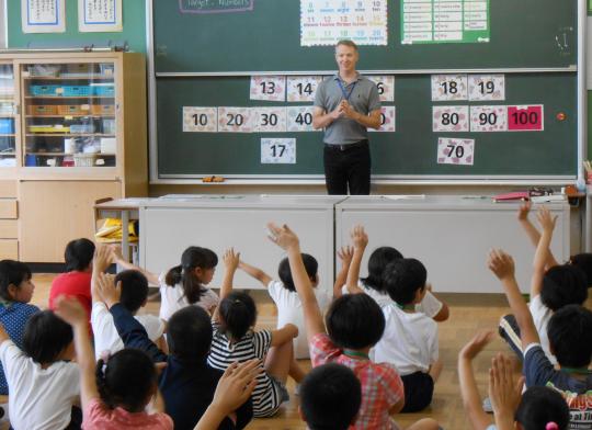 英語指導助手の男性が黒板の前に立ち、子ども達が手を上げて発言しようとしている授業風景の写真