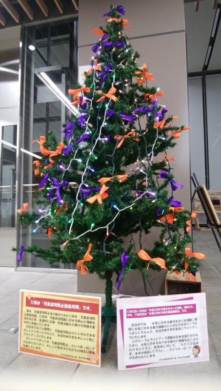 オレンジリボン・パープルリボンと電飾が飾り付けされたクリスマスツリーの下に、子どもへの虐待を防止運動と女性に対する暴力根絶運動の説明が書かれているボードが設置されている写真