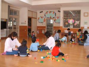 教室で先生と子ども達が床に座り、カラフルなブロックで遊んでいる写真