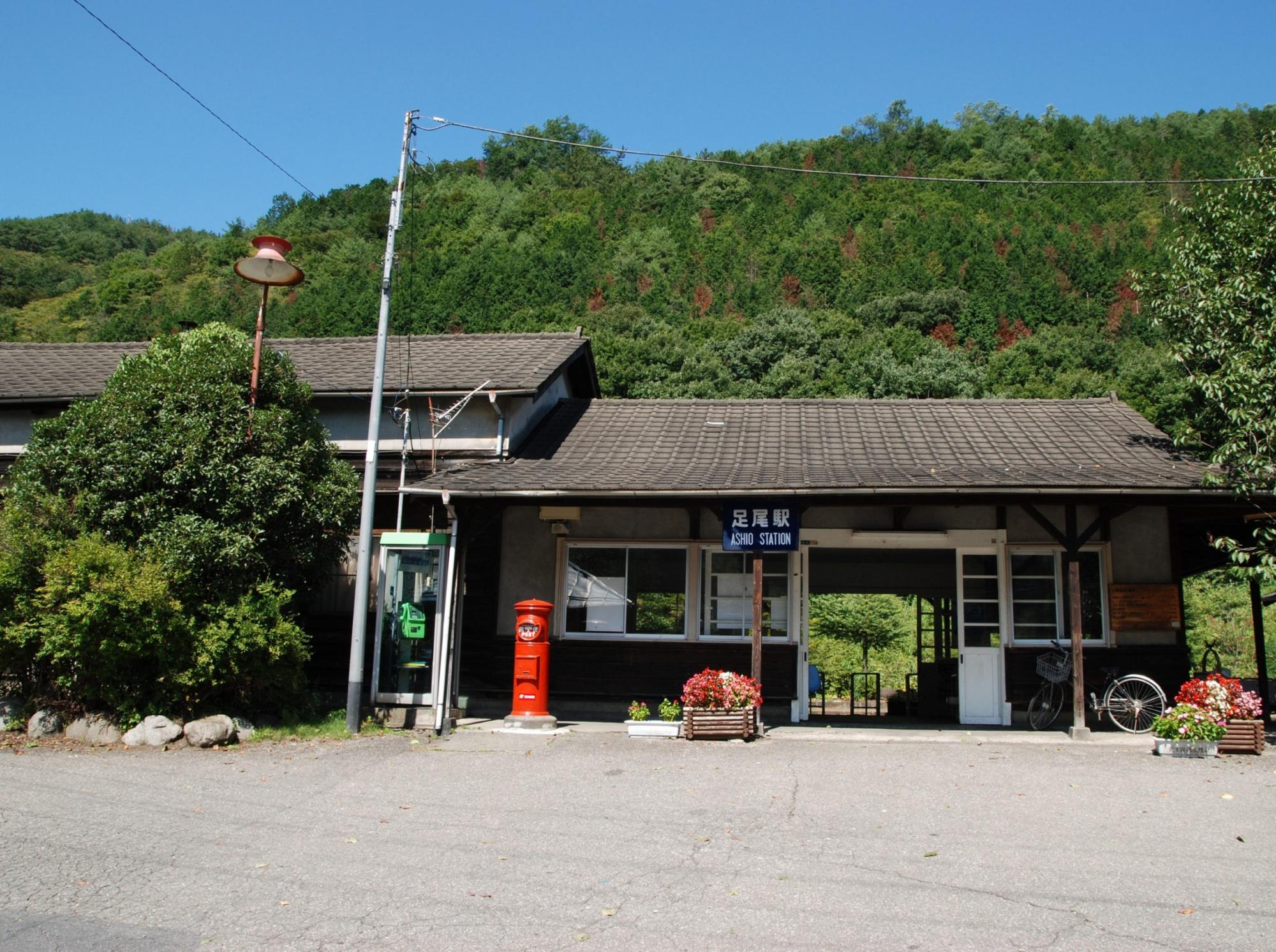 入口に赤い丸型ポストがあり、大きな緑の木の横に建っている木造でできた足尾駅の写真