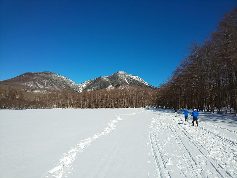 奥に山々が見え、広々とした積雪の中クロスカントリースキーをしている人2名を後方から写した写真