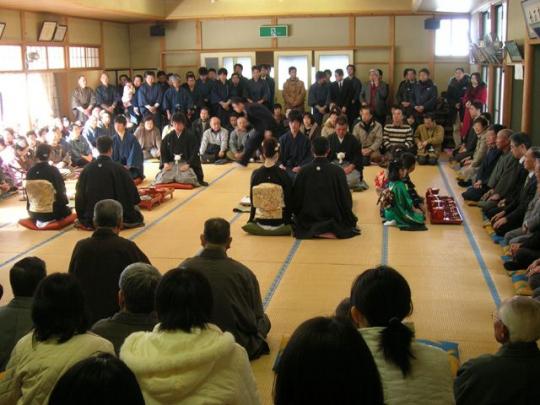 広い畳敷きの部屋で神妙に向かい合って座る着物の人たちと周りで見守る人たちの写真