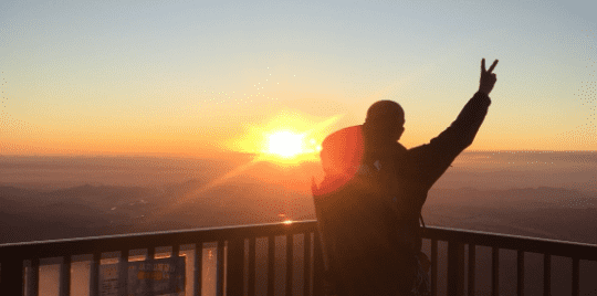 小丸山展望台で日の出をバックに右手をあげている人の写真