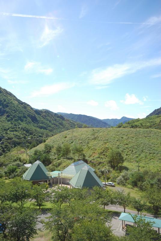 山々を背景に緑色に生い茂った木々や丘に囲まれた、ミントグリーンの三角屋根が特徴の足尾環境学習センターを丘の上から撮影した写真