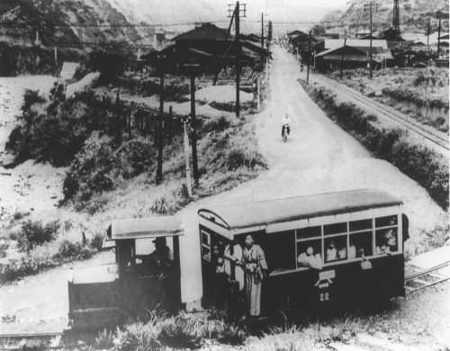 舗装がまだされていない道路と並行して線路があり、その上をバスのような見た目で運転席と乗車席が連結され、ドアや窓から乗客が身を乗り出しているガソリン気動車のモノクロ写真