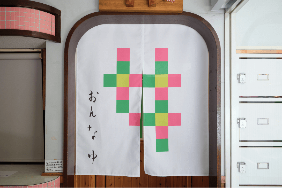 黄色、緑、ピンクの四角が集まって「女」の文字を表現し、その左横に「おんなゆ」と書かれた暖簾の写真