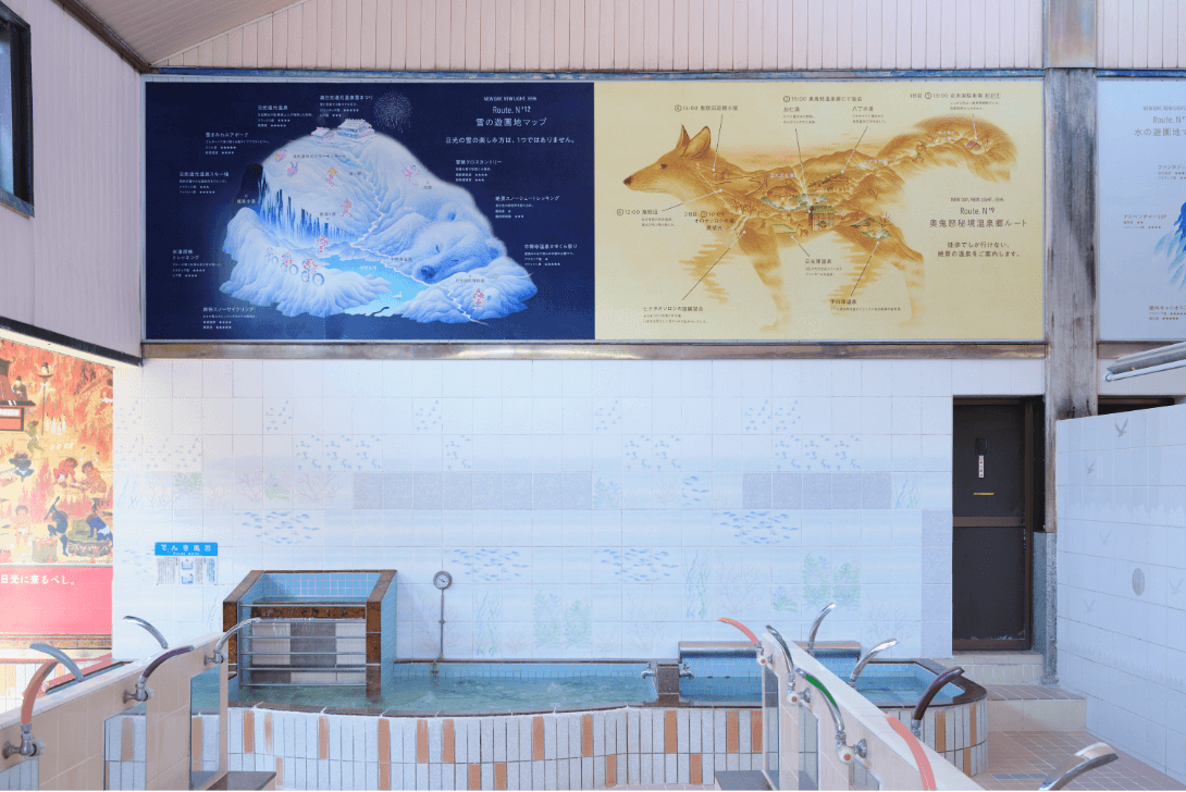 銭湯内の壁に、シロクマが寝ている姿に雪の遊園地のマップが描かれているイラストときつねを横から見た姿に奥鬼怒秘境温泉郷の場所が描かれているイラスト「NEW DAY, NEW LIGHT. 日光」のポスターが展示されている写真