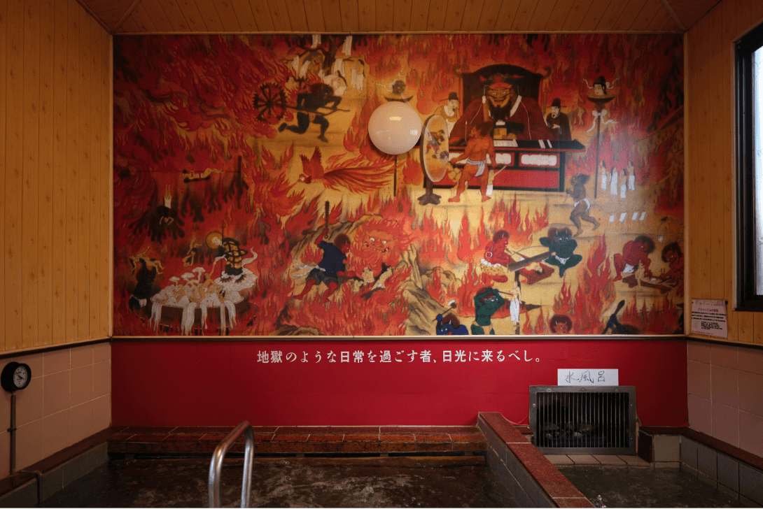 浴槽の横の壁に「地獄のような日常を過ごす者、日光に来るべし。」の文字と、炎と鬼たちのイラストが描かれている壁を写した写真