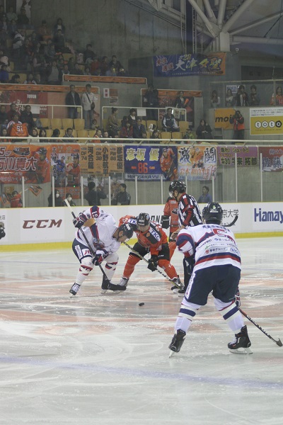 氷の上でオレンジと白いユニフォームの選手がスティックでパックの奪い合いをしているアイスバックス対イーグルスの試合の様子の写真