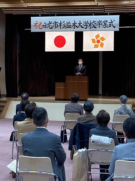 横断幕に「祝 日光市杉並木大学校卒業式」と書かれ、舞台上でスーツを着た粉川市長が挨拶するのを椅子に座り聞いている参加者の写真
