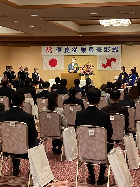「優良従業員表彰式」と書かれ、演壇で市長が話をするのを参加者が聞いている表彰式の様子の写真