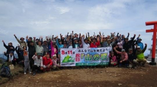 8月11日みんなで山を考えよう！祝日「山の日」と書かれた横断幕の後ろに沢山の参加者が集まり手を上に上げたポーズで写っている集合写真