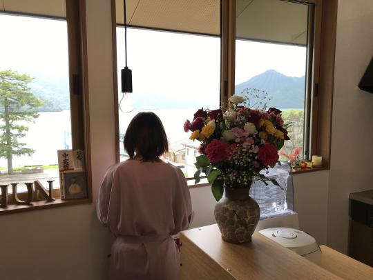 花の飾られたリビングの窓から山を望む女性の後ろ姿の写真