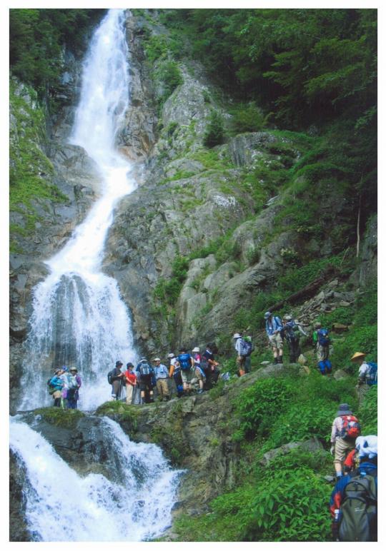 山肌を流れ落ちる赤岩滝の前を通る登山者たちの写真