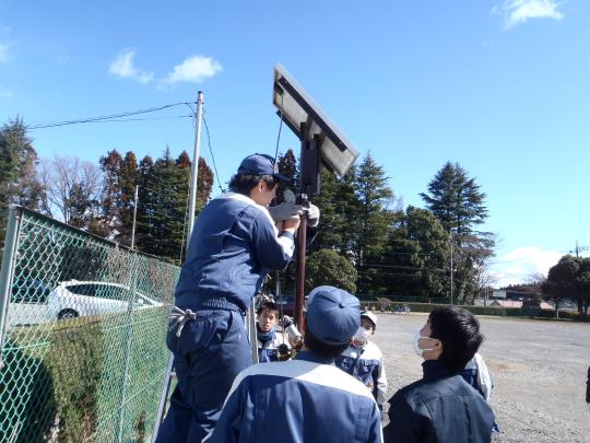 生徒たちが街路灯の柱の先端にLEDパネルをつけようとしている写真