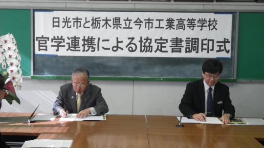 「日光市と栃木県立今市工業高等学校学官連携による協定書調印式」と書かれた黒板の前に二人座り書面にサインしている協定式の写真