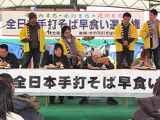 「全日本手打ちそば早食い選手権」と書かれた横断幕の前で椅子に座り手打ちそばを食べている参加者とそれを見守る関係者の写真