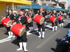 道路で法被などの祭りの衣装を着用し、太鼓を肩に掛け演奏をしている六斎市参加者の写真