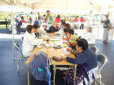 テントが張られた屋外で、長テーブルと椅子が置かれ老若男女の参加者が座って飲食をしている様子の写真