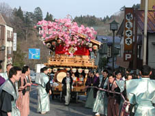 街中を大勢の袴を着用した男性が艶やかな飾りが施された神輿を綱で引いて移動している弥生祭の写真