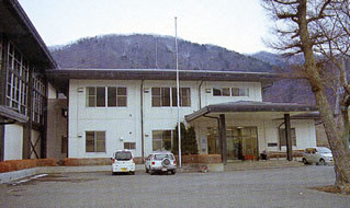 山々を背景に、白色を基調とした外壁で2階建ての玄関入口の両隣に駐車スペースが設けられている女性サポートセンター外観写真