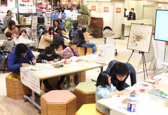 商業施設の一角で長テーブルと椅子が置かれ、大勢の親子が紙に絵を描いているワークショップの様子を写した写真