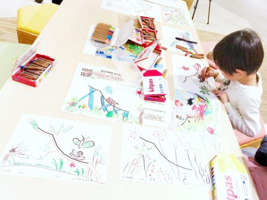 長テーブルにクレヨンや色彩豊かに描かれた作品が置かれており、そのテーブルで男の子が絵を描いている写真