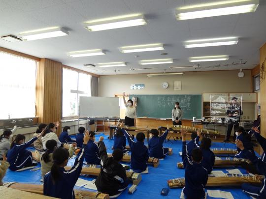 教室の前方で手を挙げている女性講師と、箏を置き床に座って手を挙げている子ども達の写真