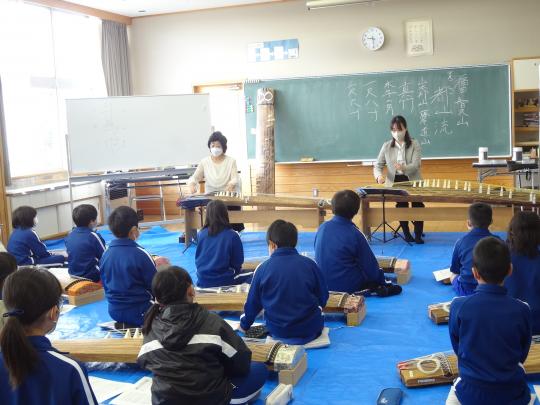 教室の前方で2人の女性講師が箏の手本を見せ、箏を置き床に座って注目している子ども達の写真