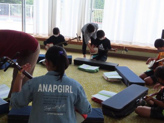 大きな窓がある室内で、床に座って三味線を弾いている子ども達の指導をしている講師の様子の写真