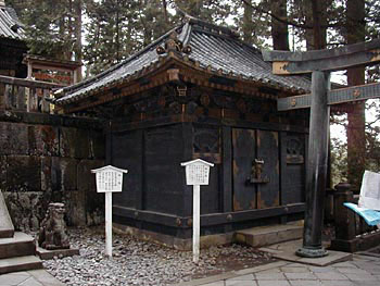 向かって神庫左側に石造りの狛犬、右側に鳥居の一部が見え、中央にある全体が黒色で寄棟造の奥社銅神庫を写した写真